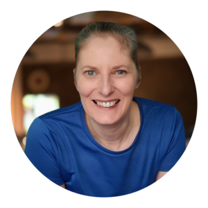 Image of a smiling white woman looking directly at the camera with their hair tied back, wearing a royal blue tshirt, set against a softly blurred indoor background.