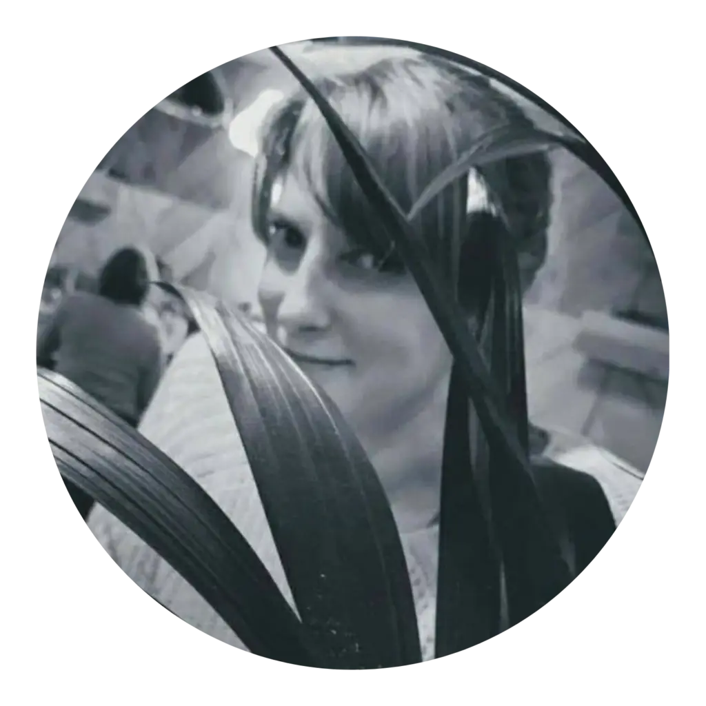 Black and white photo of Emma (a white woman with blonde hair and blue eyes) partially hidden behind the leaves of a plant, smiling slightly towards the camera. The setting appears to be indoors is in a restaurant, with blurred figures and ambient lighting in the background.