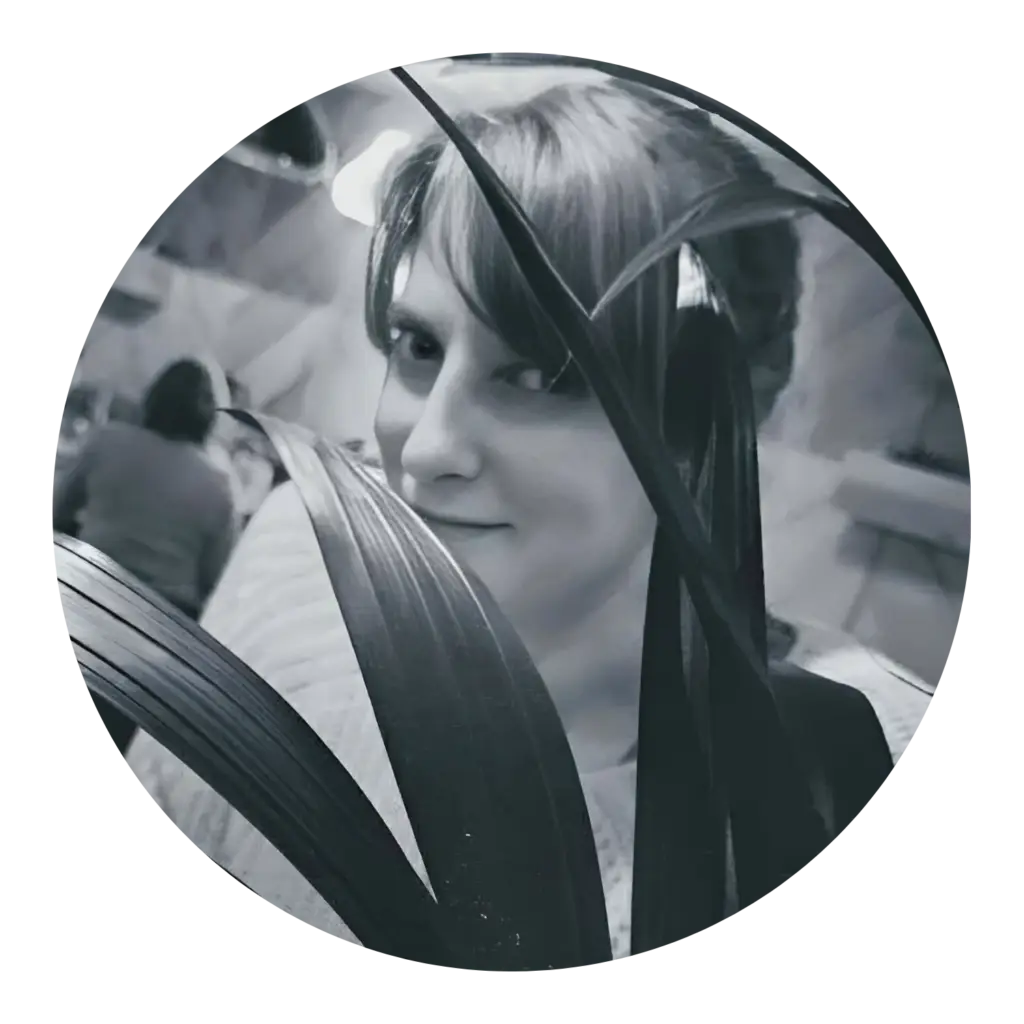 Virtual Assistant for Small Businesses. Black and white photo of Emma (a white woman with blonde hair and blue eyes) partially hidden behind the leaves of a plant, smiling slightly towards the camera. The setting appears to be indoors is in a restaurant, with blurred figures and ambient lighting in the background.