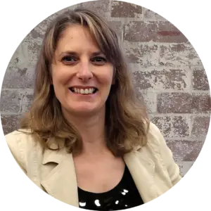 Admin Corner coworking sessions. Image of Emma (blonde white female) smiling into the camera wearing a black top and cream jack, with a rustic brick wall background.