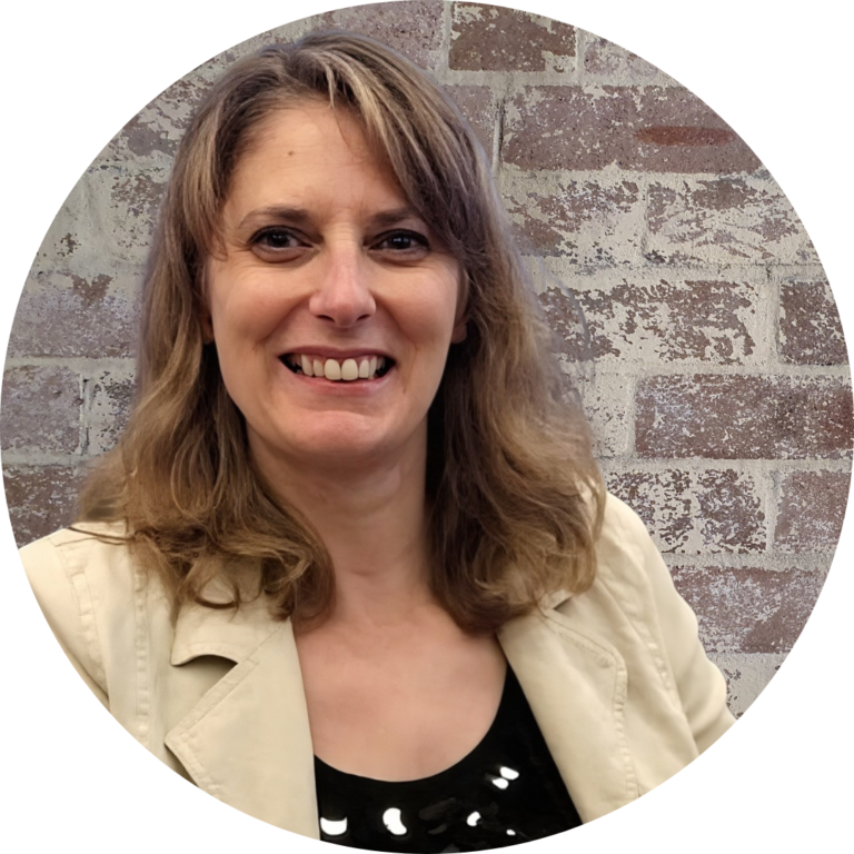 About admin consultancy. Image of Emma (blonde white female) smiling into the camera wearing a black top and cream jack, with a rustic brick wall background.